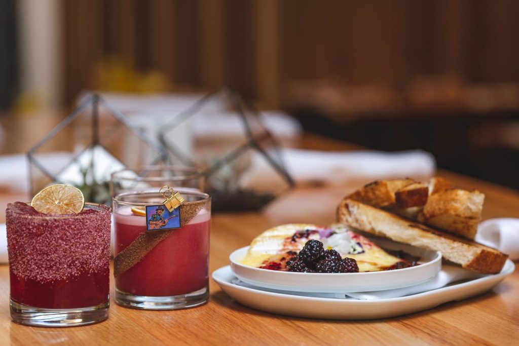Gorgeous plate of baked brie topped with blackberries beside dark raspberry colored cocktails served at Perch + Plow